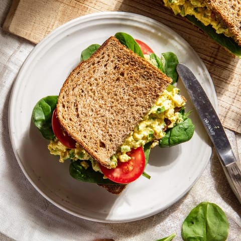 Golden avocado egg salad sandwich on toasted whole grain bread with fresh spinach and sliced tomatoes, topped with chives.  