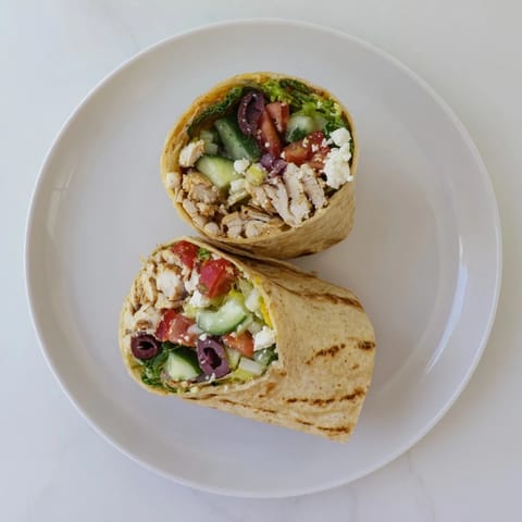 A close-up of a Greek Lemon Chicken Salad Wrap, with juicy lemon-marinated chicken, chopped cucumbers, tomatoes, and crumbled feta cheese spilling from the warm tortilla.